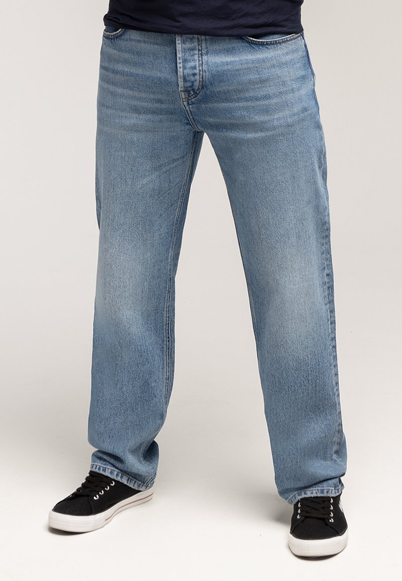 Jasnoniebieskie dżinsy z denimu o luźnym kroju, prostych nogawkach, detalach postarzenia i klasycznym pięciokieszeniowym fasonie, zestawione z czarnymi trampkami.