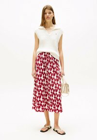 Witte gebreide mouwloze top met een polokraag, gecombineerd met een rood en wit bloemen midi-jurk. Model houdt een kleine beige handtas vast.