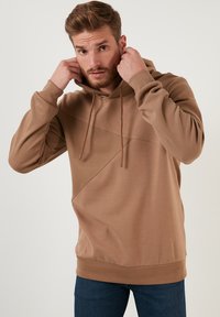 Sudadera marrón con una textura acanalada, que presenta un detalle de costura diagonal, capucha con cordón y un corte relajado. Combinada con unos jeans azules.