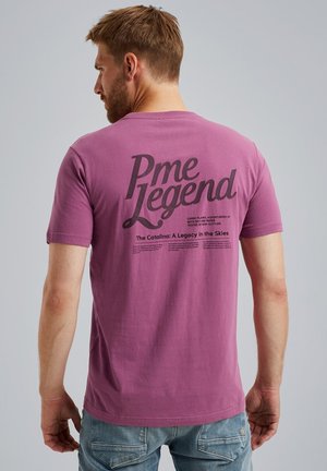 Lila bomba majica z velikim črnim napisom "Pme Legend" na hrbtu, z dodatnim besedilom pod njim. Oblika s kratkimi rokavi in okroglim izrezom.