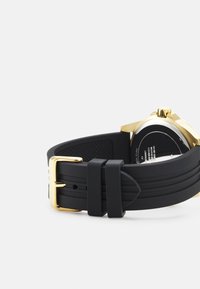 Bracelet de montre en silicone noir avec une boucle en métal doré ; texture lisse et ridules subtiles. L'arrière présente un cadran rond avec des détails mécaniques visibles.