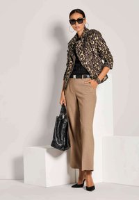 Vrouw in een panterprint jas, zwarte top, beige broek, zwarte hakken, grote zwarte handtas, creolen en zonnebril, die binnen poseert.