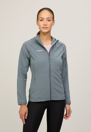 Kvinde iført en grå Mammut lynlås-hættetrøje og sorte leggings, stående mod en ensfarvet lys baggrund.