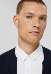 Navy cardigan over a white collared shirt, featuring a smooth texture and subtle button detail, with a close-up view of facial features.