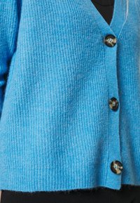 Cardigan azul claro con textura de canalé, con escote en V, cinco grandes botones oscuros y un diseño cropped. Material de lana suave.