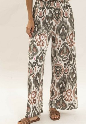 Femme portant un pantalon taille haute à jambes larges avec un motif abstrait marron, noir et beige, associé à des sandales ouvertes marron.