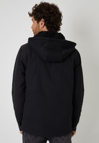 Czarna kurtka z kapturem, z teksturowanym podbiciem z sztucznego futra i zapinanymi mankietami. Projekt jest prosty, z luźnym krojem i gładkim materiałem.