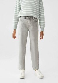 Person som bär ljusgrå högmidjade jeans, vita sneakers och en mintgrön och vit randig långärmad tröja, stående mot en vit bakgrund.