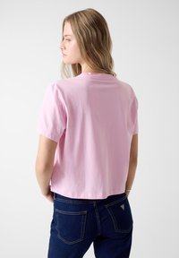 Różowy T-shirt z krótkim rękawem i okrągłym dekoltem, wykonany z bawełny, o kroju skróconym i gładkiej fakturze. Noszony z niebieskimi dżinsami.