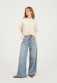 Crème korte cardigan van zacht materiaal, met knopen en twee voorkruisen, gedragen met lichtblauwe wijdlopende jeans.