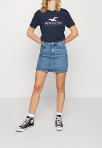 Camiseta navy con el logo de "Hollister California", combinada con una falda corta de mezclilla azul claro y zapatillas negras altas con detalles en blanco.