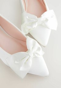 Chaussures plates en satin blanc avec un bout pointu, ornées de grands nœuds blancs à l'avant, et d'une doublure rose. Texture lisse sur l'ensemble.