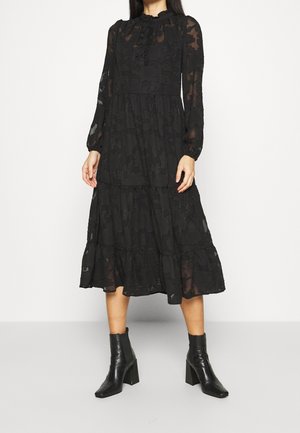 Femme portant une robe midi noire à manches longues avec texture florale et des bottines noires à talons sur un fond uni.