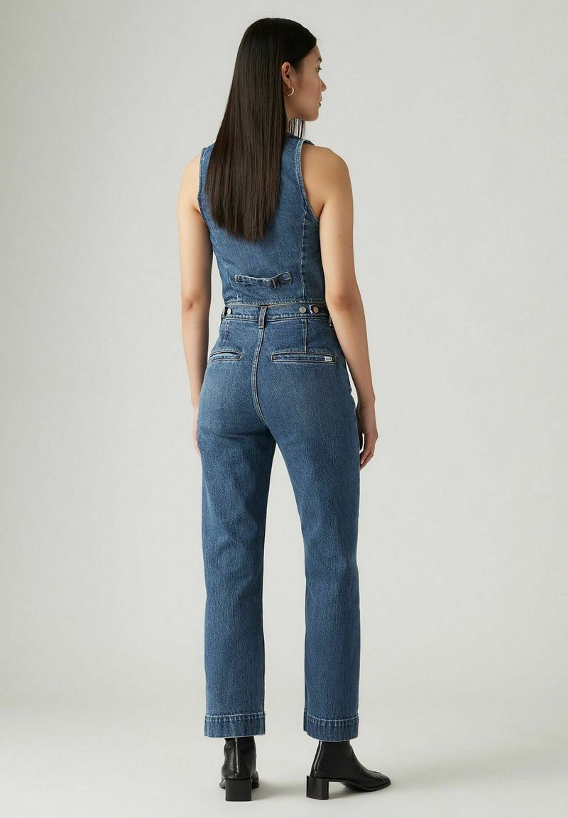 Denim-Jumpsuit in Dunkelblau, ohne Ärmel, mit Knopfverschluss hinten und schmal zulaufenden Beinen mit umgeschlagenen Säumen. Schwarze Knöchelstiefel.