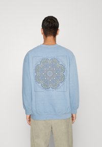 Blå sweatshirt med en rund, färgglad mandala-design på baksidan. Har ribbade ärmslut och fåll, samt en avslappnad passform.