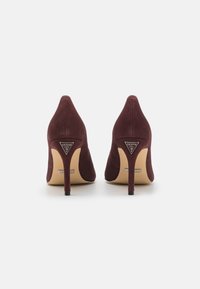 Bordo zomšos aukštakulniai su smailiais pirštais, lieknais stiletto kulnais ir metaliniais prekės ženklo akcentais nugaroje. Lygus paviršius, pakeltas dizainas.