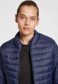Giacca trapuntata blu navy con colletto alto, caratterizzata da cuciture a righe orizzontali e una zip frontale. Indossata sopra una camicia nera.