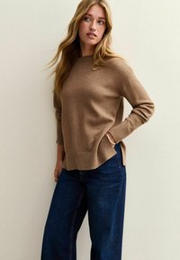 Brun stickad tröja med rund halsringning, avslappnad passform, sidofickor, i kombination med mörkblå vida jeans, mot en ljus bakgrund.