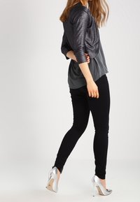 Zapatos de tacón alto plateados, jeans ajustados negros y una parte superior gris oscura con una chaqueta corta negra brillante, que muestra un diseño en capas contrastante.