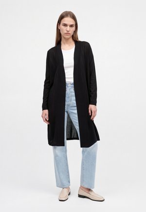 Le modèle se tient devant un fond de studio blanc, vêtu d’un long cardigan noir, d’un haut blanc, d’un jean bleu clair et de mocassins beiges.
