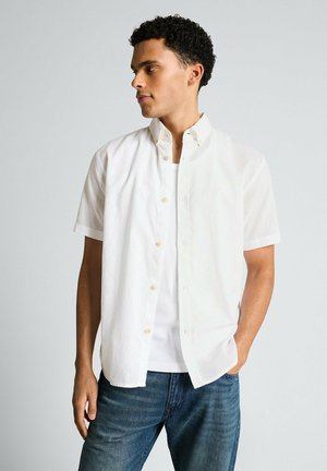 Mężczyzna z kręconymi włosami, ubrany w białą koszulę z krótkim rękawem i guzikami na białym tank topie oraz w niebieskie dżinsy, patrzący w lewo.