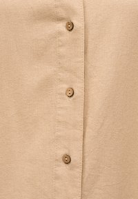 Μπεζ υφασμάτινο πουκάμισο μπροστινή όψη σε κοντινό πλάνο που δείχνει τρία μικρά στρογγυλά ξύλινα κουμπιά σε ραμμένη πατιλέτα.