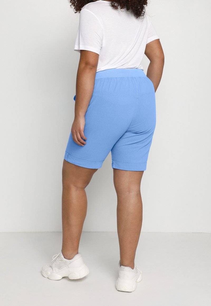 Personne portant un short bleu clair jusqu'aux genoux, une chemise blanche à manches courtes et des baskets blanches, debout devant un fond uni gris clair.