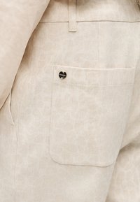 Κοντινό πλάνο μπεζ υφασμάτινων παντελονιών με ορθογώνια πλαϊνή τσέπη και ένα μικρό στρογγυλό μαύρο και χρυσό μεταλλικό κουμπί με λογότυπο.