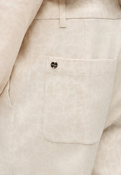 Gros plan sur un pantalon en tissu beige avec une poche latérale rectangulaire ornée d'un petit bouton métallique rond noir et doré à logo.