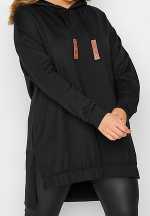 Czarna, oversize bluza z kapturem z brązowymi troczkami, noszona z czarnymi legginsami, ręce splecione z przodu, przycięta na wysokości ramion i śródudzia.