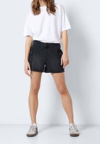 Schwarze Denim-Cargo-Shorts mit zwei seitlichen Taschen, hoher Taille und lockerer Passform, kombiniert mit einem weißen, übergroßen T-Shirt und grauen Turnschuhen.