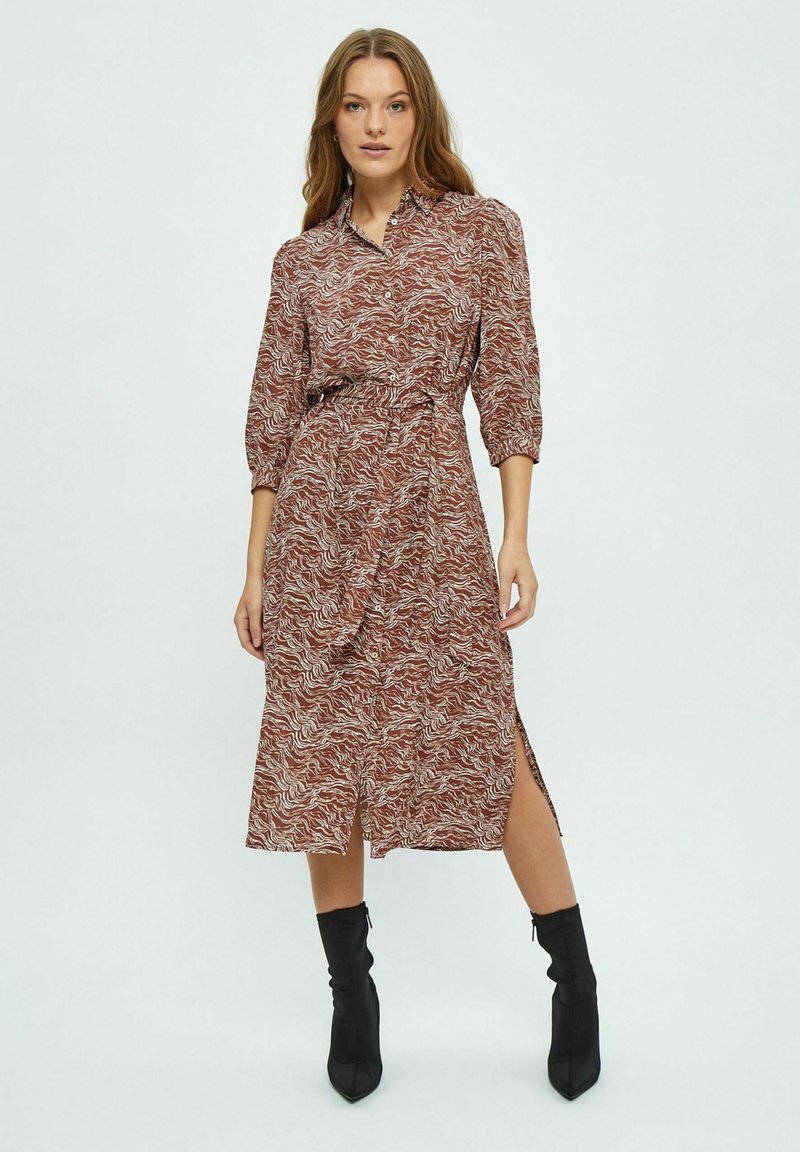 Braunes gemustertes Kleid mit durchgehender Knopfleiste, Dreiviertelärmeln, elastischem Taillenbund und seitlichen Schlitzen, kombiniert mit schwarzen Knöchelstiefeln.