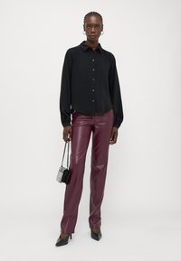 Camisa preta de botão com mangas longas, combinada com calças em imitação de pele borgonha. O modelo segura uma pequena bolsa preta e usa saltos pretos.