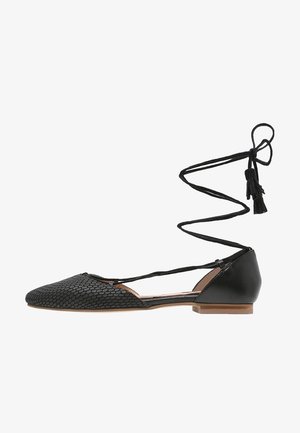 Scarpa nera piatta con punta appuntita texturizzata, lacci avvolgenti intorno alla caviglia con nappine e controcanto liscio con suola in legno.