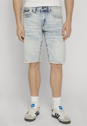 Jasnoniebieskie jeansowe szorty z postrzępionymi brzegami, noszone przez osobę w białych skarpetkach i trampkach z czarnymi paskami.