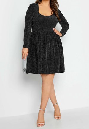 Femme portant une robe noire longue à manches longues scintillante, associée à des sandales argentées à talons hauts et tenant une pochette argentée.