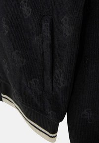 Veste noire côtelée avec un motif de logo discret, poche latérale et bande élastique rayée à l'ourlet inférieur. Tissu texturé avec des détails tricotés.