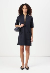 Navyblaues Kurzarmkleid mit einem Reißverschluss am Kragen, zwei vorderen Taschen und lockerer Passform. Kombiniert mit einer kleinen schwarzen Tasche und flachen Schuhen.