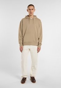 Felpa beige con tasca a marsupio frontale, cappuccio regolabile con cordino e dettaglio del logo. Abbinata a pantaloni bianchi e scarpe marroni.
