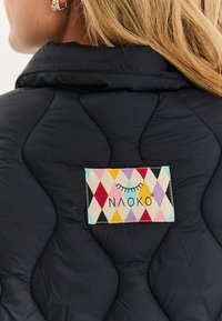 Blond osoba v černé prošívané bundě s barevnou nášivkou značky Naoko ve tvaru diamantů na horní části zad pod límcem.