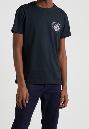 Homme portant un t-shirt noir Versace Jeans avec un logo tigre sur la poitrine et un jean bleu foncé, debout devant un fond clair uni.