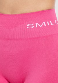Leggings rosa senza cuciture in tessuto elasticizzato con vita alta, decorata con il nome del marchio "SMILOS". Texture liscia e design aderente.