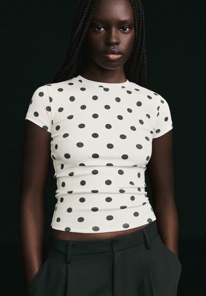 Kobieta z długimi zaplecionymi włosami, ubrana w dopasowaną białą bluzkę z krótkim rękawem w czarne kropki, w połączeniu z czarnymi spodniami, na ciemnym tle.