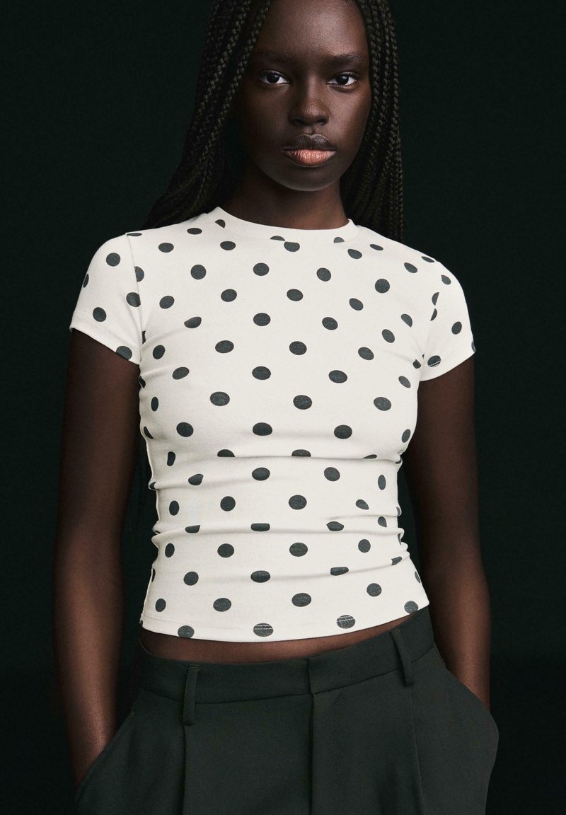 Femme aux longs cheveux tressés portant un haut blanc ajusté à manches courtes avec des pois noirs, associée à un pantalon noir, sur un fond sombre.