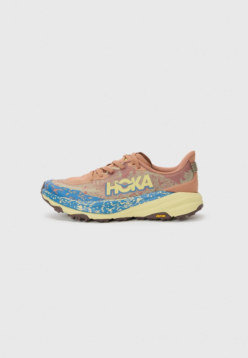 Trailrunning-Schuh mit einem Pfirsichfarbigen Obermaterial, blauen und gelben Designelementen, strukturiertem Sohlenprofil und Vibram-Logo auf der Außensohle.