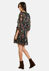 Kobieta w przezroczystej czarnej sukience w kwiaty z marszczoną talią i rękawami trzyczwartowymi, w ciemnobrązowych botkach na obcasie, idąca w lewo.