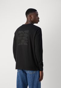 Černé tričko s dlouhým rukávem vyrobené z hladkého materiálu, s velkým černým grafickým motivem na zadní straně. Ležérní design s kulatým výstřihem a přiléhavými rukávy.