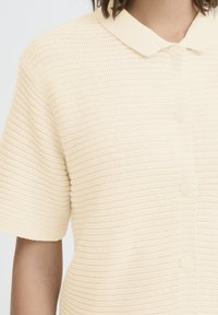 Un gilet en tricot à manches courtes de couleur crème avec un motif de points texturé, un col rond et des boutons assortis sur le devant, porté par une personne.