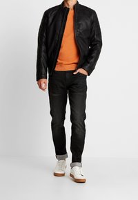 Veste en cuir noire avec des détails aux épaules, pull orange, jeans noirs et baskets blanches avec des semelles brunes. Fond uni.