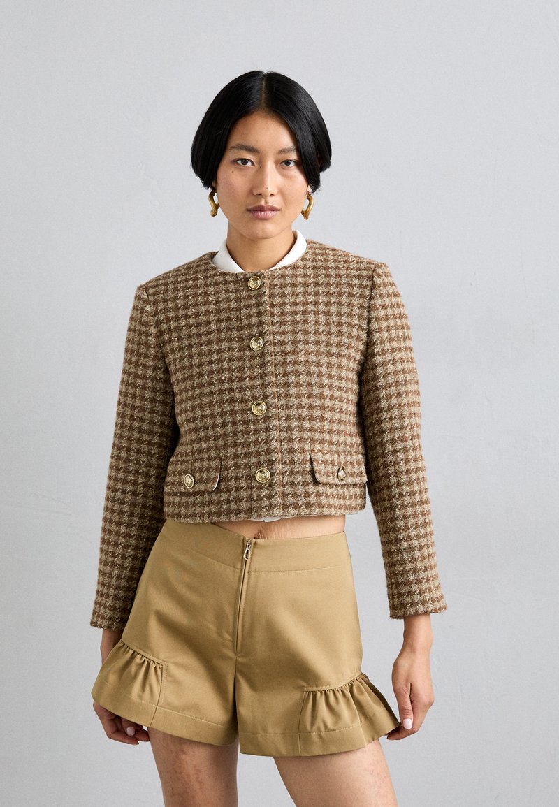Chaqueta cropped marrón de patrón houndstooth con botones dorados y bolsillos, combinada con pantalones cortos caqui que tienen un dobladillo con volantes y cremallera en la parte trasera.